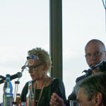 Drei Personen bei Lesung im 26. Stock: Frau am Mikrofon mit Brille, Mann rechts, moderierter Event mit Publikum