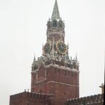Die Spasskaja-Turm-Uhr des Moskauer Kremls im Winter: Der markante rote Backsteinturm mit goldenen Zifferblättern, bekrönt von einem roten Stern, ragt vor einem grauen, verschneiten Himmel. Die Mauern des Kremls und schneebedeckte Bäume im Vordergrund vermitteln eine winterliche, historische Atmosphäre.