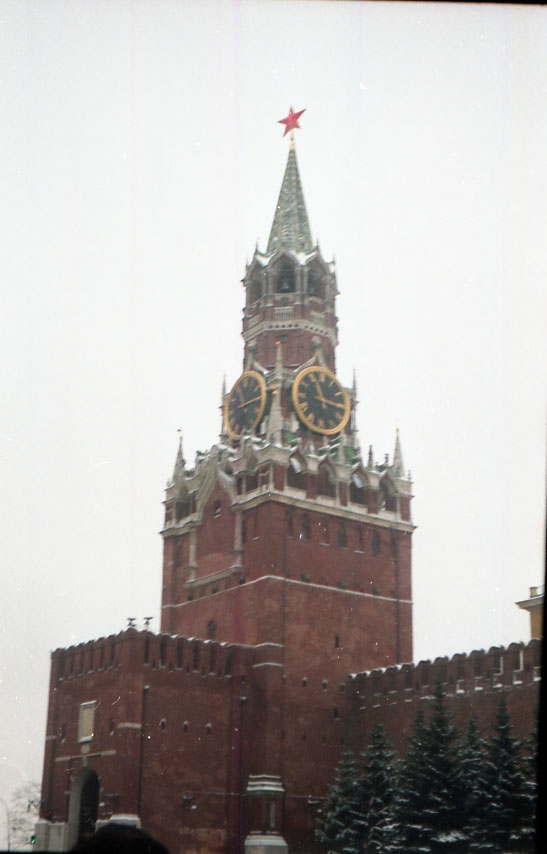 Die Spasskaja-Turm-Uhr des Moskauer Kremls im Winter: Der markante rote Backsteinturm mit goldenen Zifferblättern, bekrönt von einem roten Stern, ragt vor einem grauen, verschneiten Himmel. Die Mauern des Kremls und schneebedeckte Bäume im Vordergrund vermitteln eine winterliche, historische Atmosphäre.