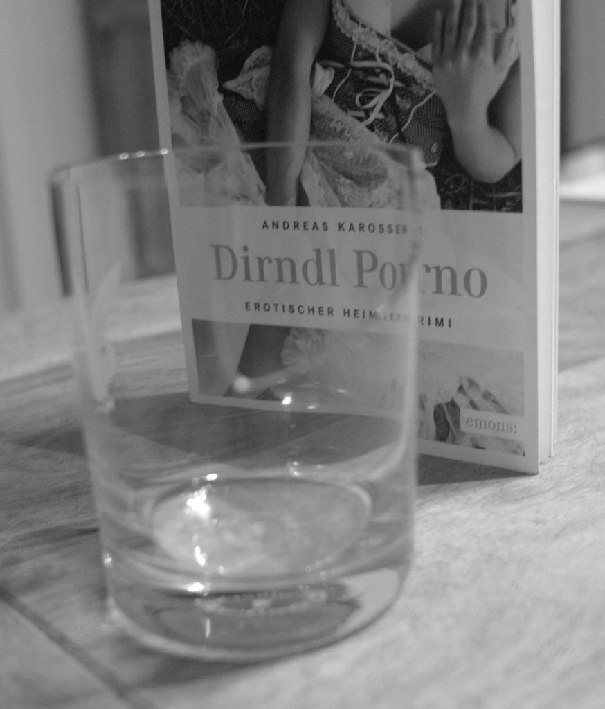 Buch "Dirndl Porno" von Andreas Karosser auf Tisch neben Wasserglas, Schwarzweiß-Fotografie