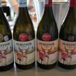 Vier Flaschen Survivor Wein aus Swartland Südafrika nebeneinander, jede mit einer roten Kuh auf dem Etikett, daneben ein W…