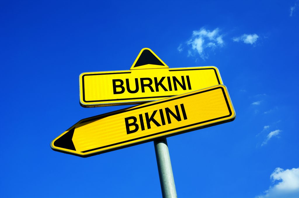 Gelbes Wegweiserschild mit Richtungspfeilen für "Burkini" und "Bikini" vor blauem Himmel