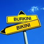 Gelbes Wegweiserschild mit Richtungspfeilen für "Burkini" und "Bikini" vor blauem Himmel