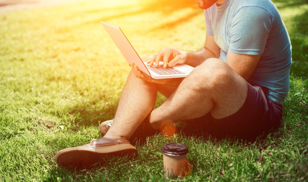 Mann sitzt auf Rasen mit Laptop und Kaffee im Freien beim Homeoffice arbeiten