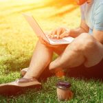 Mann sitzt auf Rasen mit Laptop und Kaffee im Freien beim Homeoffice arbeiten