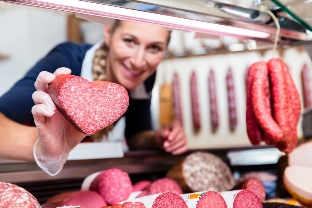 Fleischverkäuferin in Metzgerei hält herzförmige Wurst in die Kamera, dahinter Wurstausstellung und Regale mit Fleischwaren