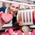Fleischverkäuferin in Metzgerei hält herzförmige Wurst in die Kamera, dahinter Wurstausstellung und Regale mit Fleischwaren