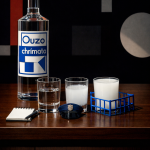 Stillleben auf einem Holztisch: Eine klare Ouzo-Flasche mit blau-weißem Etikett steht links. Daneben drei Gläser – eines mit klarem Ouzo, eines mit milchig getrübtem Ouzo nach Wasserzugabe und eines vollständig weiß. Vor den Gläsern liegen ein kleines Notizbuch mit Stift und eine blaue Polizeimütze. Rechts steht ein Glas in einem blauen Drahtkorb. Der dunkle Hintergrund zeigt abstrakte geometrische Formen.
