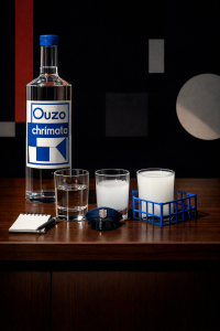 Stillleben auf einem Holztisch: Eine klare Ouzo-Flasche mit blau-weißem Etikett steht links. Daneben drei Gläser – eines mit klarem Ouzo, eines mit milchig getrübtem Ouzo nach Wasserzugabe und eines vollständig weiß. Vor den Gläsern liegen ein kleines Notizbuch mit Stift und eine blaue Polizeimütze. Rechts steht ein Glas in einem blauen Drahtkorb. Der dunkle Hintergrund zeigt abstrakte geometrische Formen.