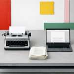 Ein minimalistischer Schreibtisch mit einer klassischen mechanischen Schreibmaschine (links) und einem modernen Laptop (rechts), dazwischen ein Stapel bedruckter Manuskriptseiten. Im Hintergrund eine weiße Wand mit farbigen geometrischen Flächen in Rot, Gelb, Grün und Rosa. Das Bild symbolisiert den Übergang von traditionellem zu digitalem Schreiben