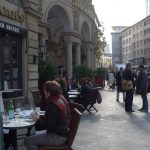 Straßenszene in Frankfurt vor dem historischen Gebäude des Steigenberger Frankfurter Hof: Menschen sitzen an kleinen runden Tischen eines Straßencafés, trinken Kaffee und unterhalten sich, während im Hintergrund Geschäftsleute auf dem Gehweg stehen und sprechen. Die Nachmittagssonne fällt zwischen modernen Hochhäusern und Altbaufassade auf die belebte Innenstadt
