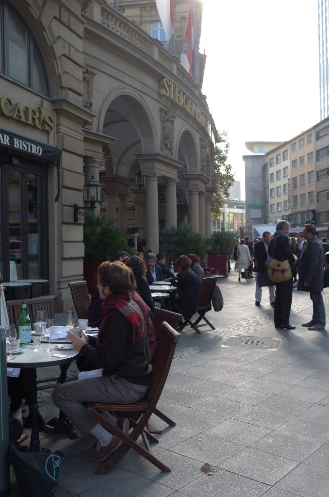 Straßenszene in Frankfurt vor dem historischen Gebäude des Steigenberger Frankfurter Hof: Menschen sitzen an kleinen runden Tischen eines Straßencafés, trinken Kaffee und unterhalten sich, während im Hintergrund Geschäftsleute auf dem Gehweg stehen und sprechen. Die Nachmittagssonne fällt zwischen modernen Hochhäusern und Altbaufassade auf die belebte Innenstadt