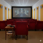 **Alt-Text für das Bild:** „Ein kleines, leeres Kino mit roten Sesseln, das auf der Leinwand eine Szene aus einem Zugabteil zeigt. Vorne steht ein Tisch mit einem Radio und Kopfhörern, darunter liegt ein Programmheft mit der Aufschrift ‚Rein Vorfilm‘. Die Wände sind in gelb-weiß gehalten, und die Atmosphäre wirkt einladend und nostalgisch.“