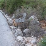 Ein verlassener, metallener Gartenstuhl steht schief zwischen wildem Gestrüpp und einer trockenen Steinmauer am Rand eines Schotterwegs in ländlicher Umgebung