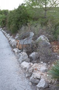 Ein verlassener, metallener Gartenstuhl steht schief zwischen wildem Gestrüpp und einer trockenen Steinmauer am Rand eines Schotterwegs in ländlicher Umgebung