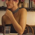 Junge Frau sitzt an einem gedeckten Restauranttisch und schaut nachdenklich zur Seite. Sie trägt ein schwarzes trägerloses Oberteil, einen breiten Strass-Choker am Hals und einen Diamant-Ohrstecker. Die Hände sind vor der Brust verschränkt, am Handgelenk ein Armreif. Auf dem Tisch stehen ein Wasserglas und Besteck. Im Hintergrund rosa Blüten und dunkle Holzregale. Warmes, gedämpftes Licht