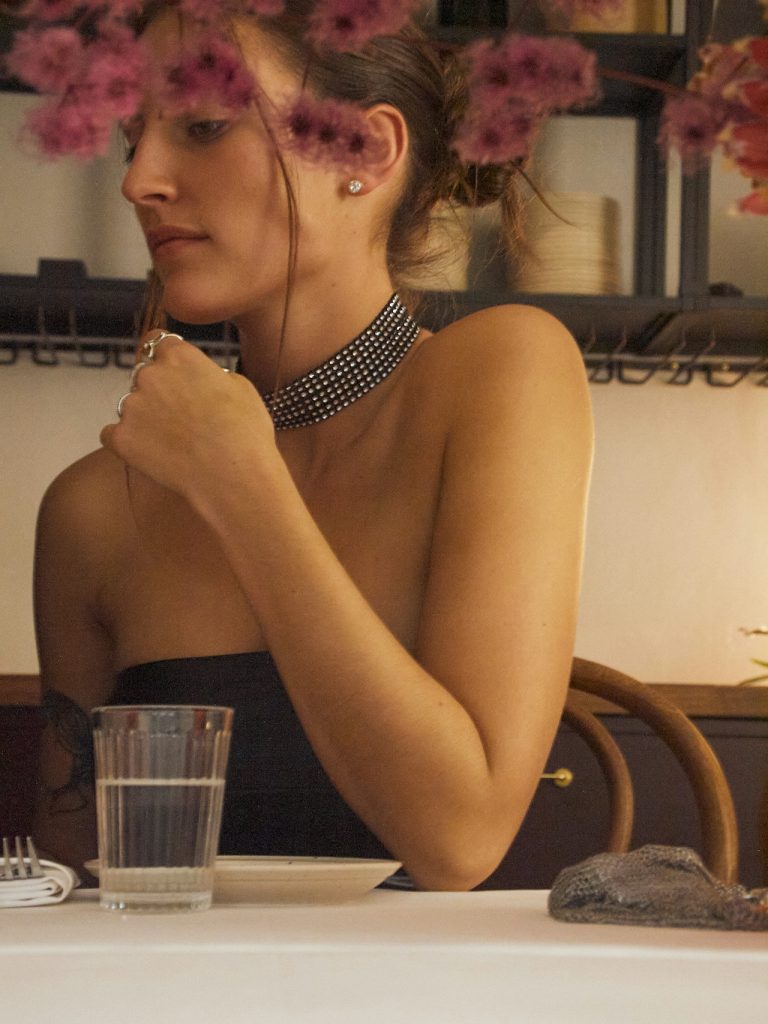 Junge Frau sitzt an einem gedeckten Restauranttisch und schaut nachdenklich zur Seite. Sie trägt ein schwarzes trägerloses Oberteil, einen breiten Strass-Choker am Hals und einen Diamant-Ohrstecker. Die Hände sind vor der Brust verschränkt, am Handgelenk ein Armreif. Auf dem Tisch stehen ein Wasserglas und Besteck. Im Hintergrund rosa Blüten und dunkle Holzregale. Warmes, gedämpftes Licht