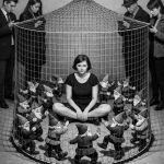 Frau sitzt in Käfig umgeben von Puppen, fünf Personen halten Netz, Schwarz-Weiß-Fotografie, surreale Künstlerische Darstel…