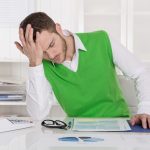 Frustrierter Mann am Schreibtisch mit Dokumenten, Brille und Taschenrechner in modernem Büro
