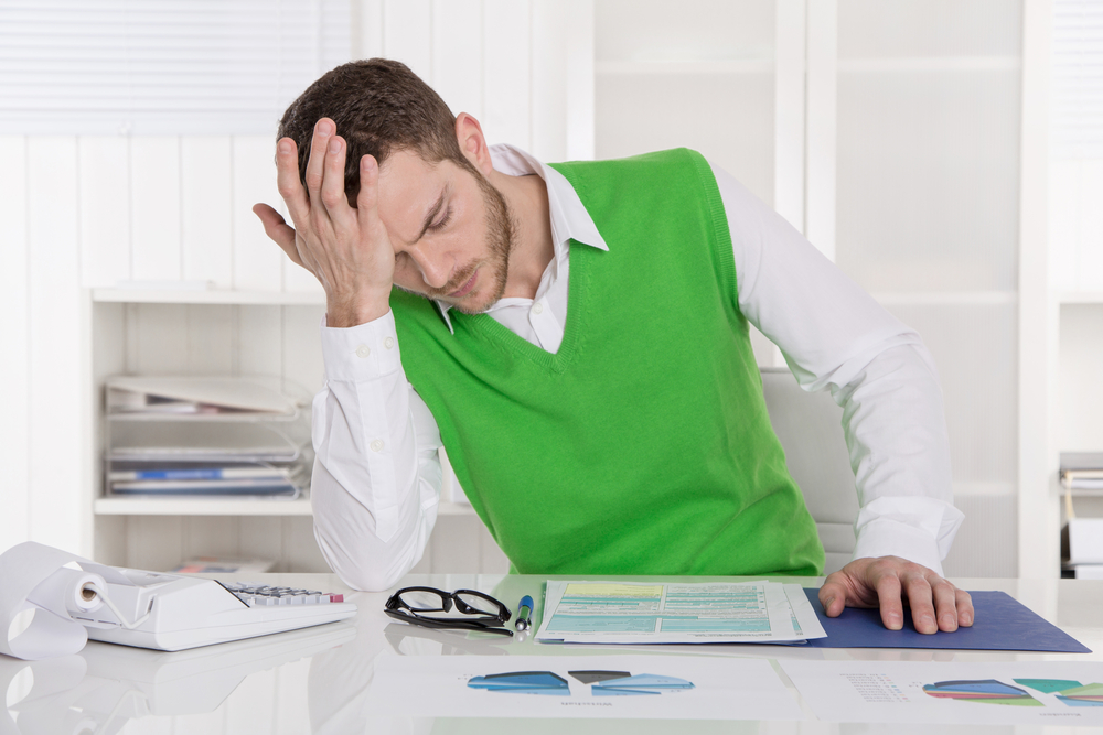 Frustrierter Mann am Schreibtisch mit Dokumenten, Brille und Taschenrechner in modernem Büro