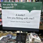 Buchhandlung-Lesezeichen mit Werbung für Dark Romance: "A knife? Are you flirting with me?" vor Bücherregal mit Romanen