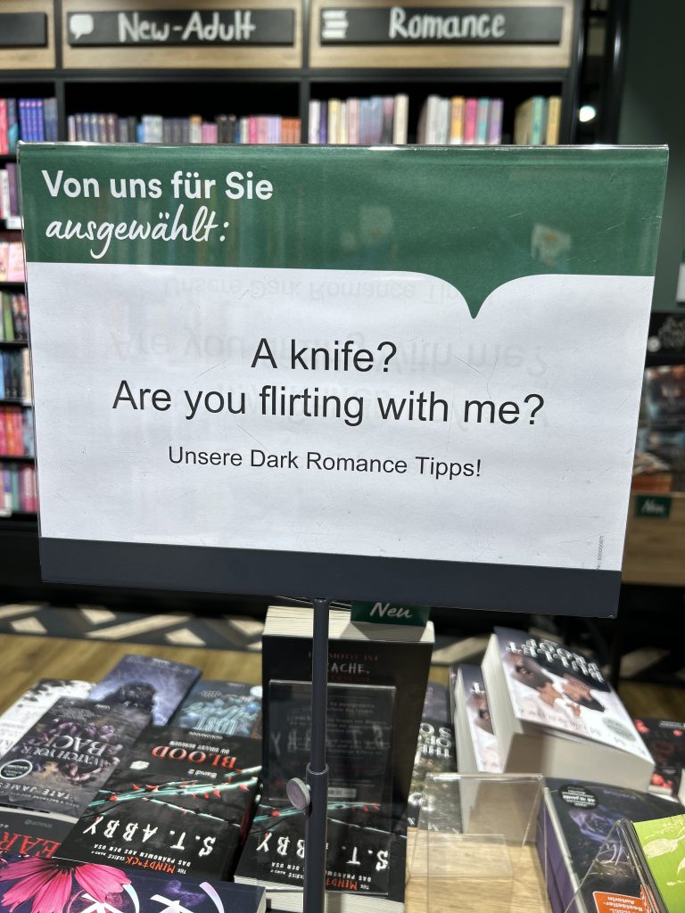 Buchhandlung-Lesezeichen mit Werbung für Dark Romance: "A knife? Are you flirting with me?" vor Bücherregal mit Romanen