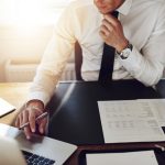 Geschäftsmann in weißem Hemd analysiert Finanzberichte und Daten am Schreibtisch mit Laptop und Kaffee