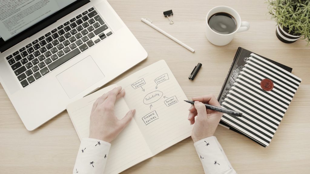 Person skizziert Produktivitäts-Diagramm am Schreibtisch mit Laptop, Kaffee und Notizbuch im modernen Büro-Workspace