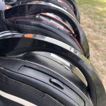 Mehrere schwarze Fahrradhelme gestapelt nebeneinander auf Gras, mit roten und weißen Akzenten und Beschriftungsetiketten