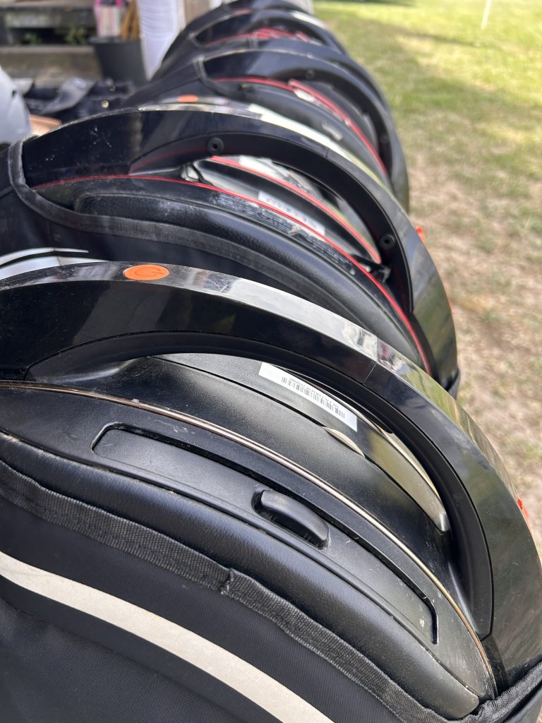 Mehrere schwarze Fahrradhelme gestapelt nebeneinander auf Gras, mit roten und weißen Akzenten und Beschriftungsetiketten
